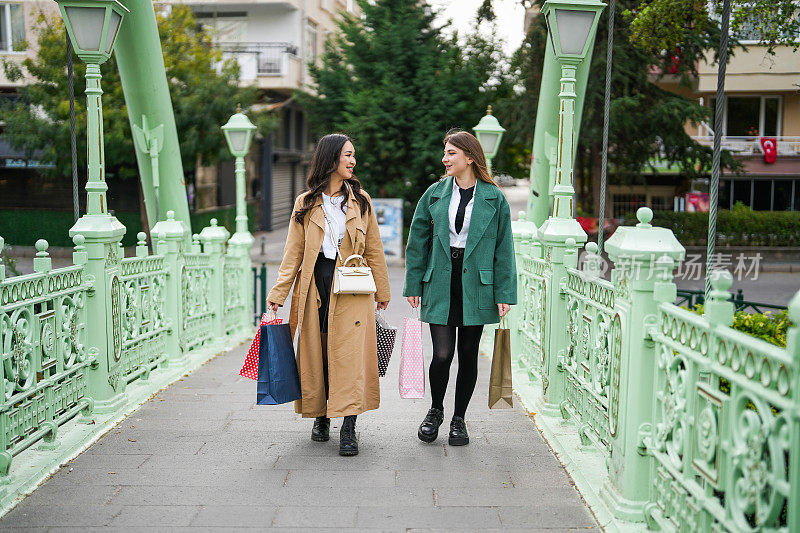 两个手里拿着购物袋的年轻女子走过市中心的一座桥。黑色星期五折扣
