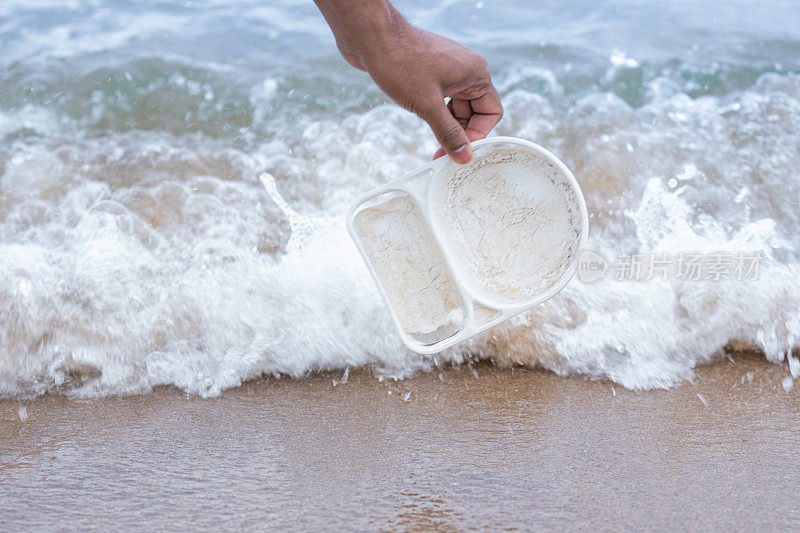 一个人的手从岸边收集塑料食品容器的特写镜头，以防止微塑料的产生。一个不知名的人拯救了塑料垃圾造成的环境。