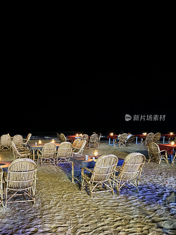 海滩餐厅夜景的图片，铺着桌布和椅子的竹制桌子，为露天用餐做好了准备，点燃的蜡烛桌子的中心，俯瞰Palolem海滩和阿拉伯海，印度果阿，黑暗的夜空背景