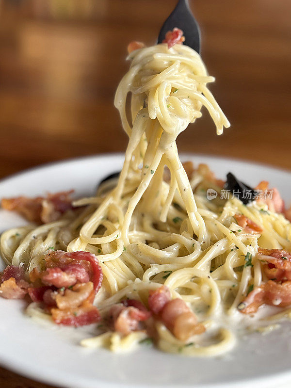 图像金属叉子旋转的意大利面条，以培根、帕尔马干酪、切碎的大蒜、鸡蛋、黑橄榄半、欧芹叶装饰在白色盘子上，木制餐厅餐桌背景，高架视图，重点放在前景上