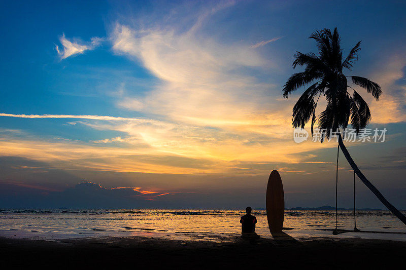 冲浪男子的剪影坐在海滩上冲浪板。冲浪场景在日落海滩与彩色的天空。户外水上运动冒险生活方式。夏天的活动。20多岁英俊的亚洲男模。