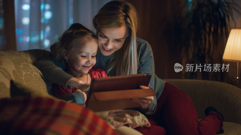 美丽的妈妈和她的小女儿正坐在客厅的沙发上，他们在用平板电脑。这是晚上，房间是舒适和温暖的。