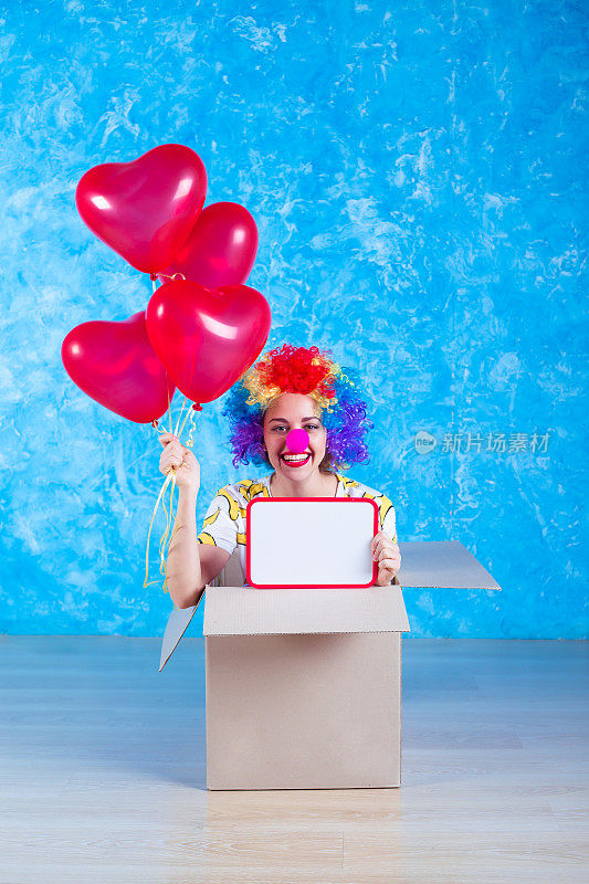 1愚人节的概念。滑稽的小丑坐在一个带着氦气球的纸箱里，看着镜头。生日礼物。