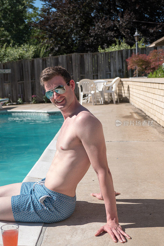 一名健康的男子坐在泳池边，脚在水里，没有穿衬衫和蓝色泳裤，戴着飞行员太阳镜看着摄像机。一个靠在泳池边的白人。