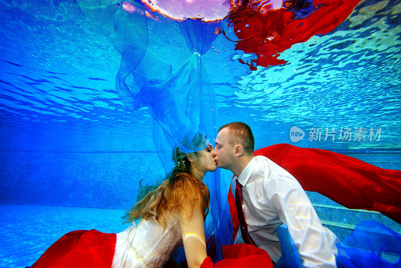 神奇的水下婚礼。幸福的新娘和新郎游泳和亲吻在夕阳下的游泳池周围的红色和蓝色织物。肖像。的概念。水下摄影