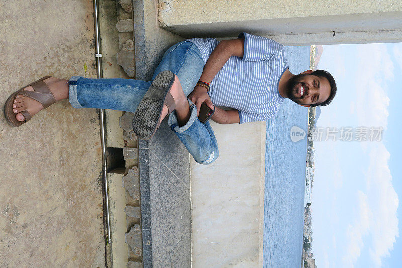 一个快乐英俊的印度男人在假日微笑，坐在阳光下的泰姬陵湖宫殿的湖景，乌代普尔，印度，年轻英俊的男人拿着手机在破蓝色牛仔裤，条纹t恤，笑完美的牙齿微笑