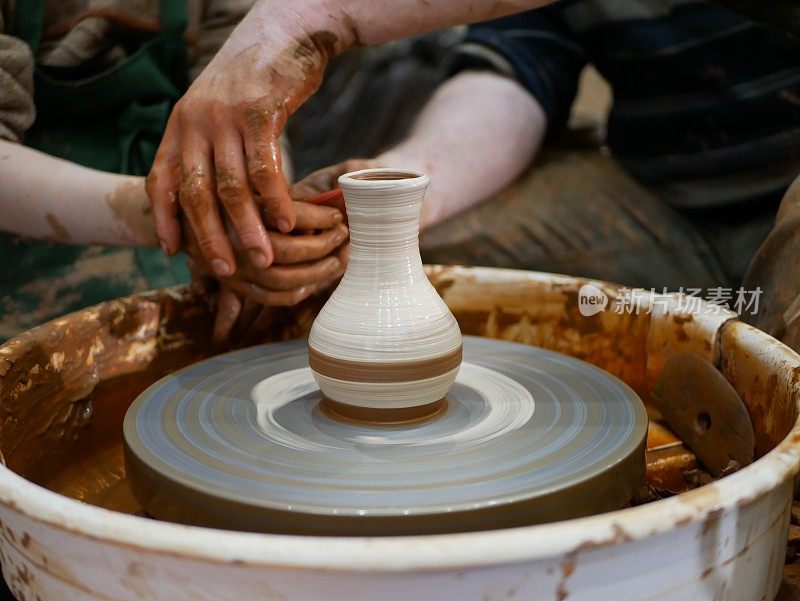 陶工师傅帮助孩子在一个电动驱动的现代陶工车轮上用白色油漆画一个陶罐。在陶瓷花瓶上画图案。一个孩子和一个成年人的手都沾满了泥土。
