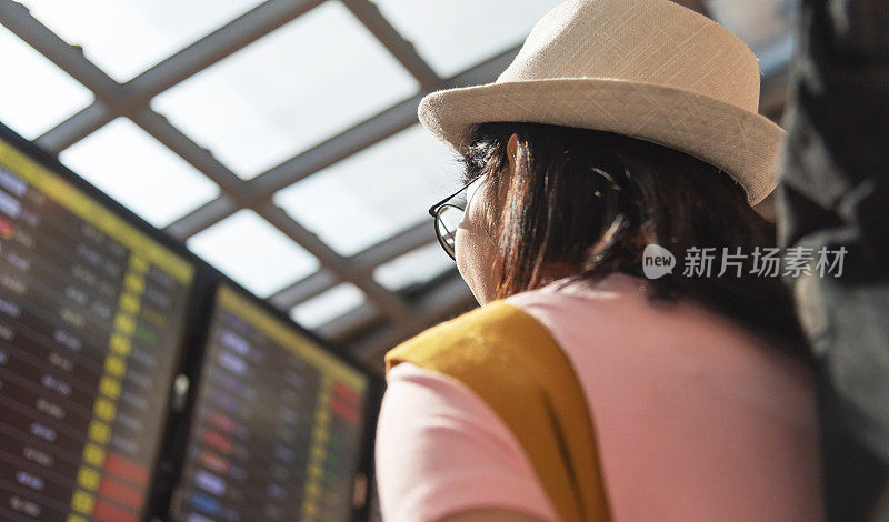 亚洲女性游客戴着帽子和粉红色的衬衫看着航班时刻表。后面的观点。