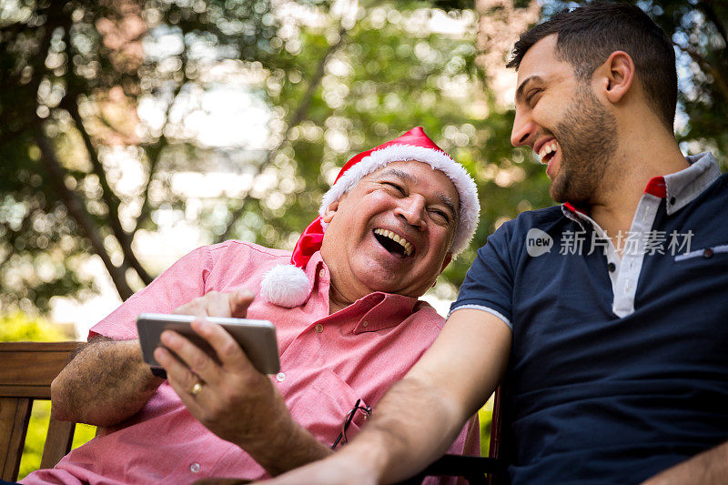 爸爸戴着圣诞老人的帽子在用智能手机，儿子在公园里玩得很开心