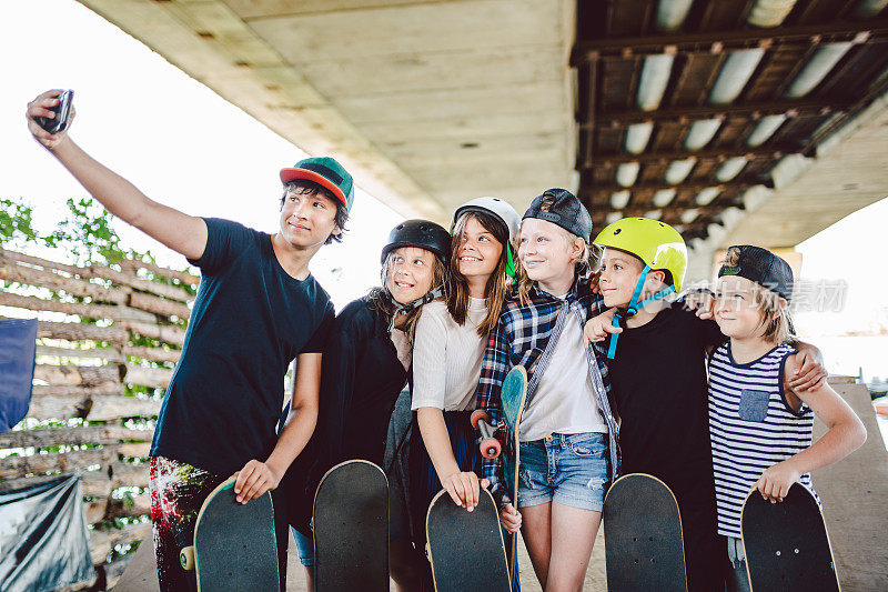 一群玩滑板的朋友在滑板公园用手机自拍。一大群带着滑板的孩子在滑板公园的坡道上拍摄回忆照片。一群孩子在智能手机上合影