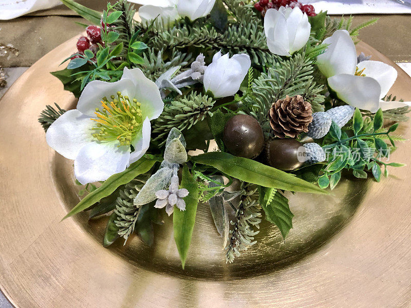 金色的盘子上装饰着人造云杉和冬青花环的圣诞餐桌，白色的圣诞玫瑰(嚏根草)花，金色的桌廊，俯瞰的景色