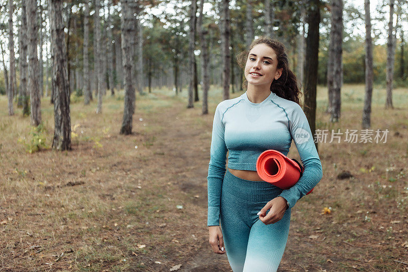 迷人的健身女人在运动服装持有瑜伽垫在森林
