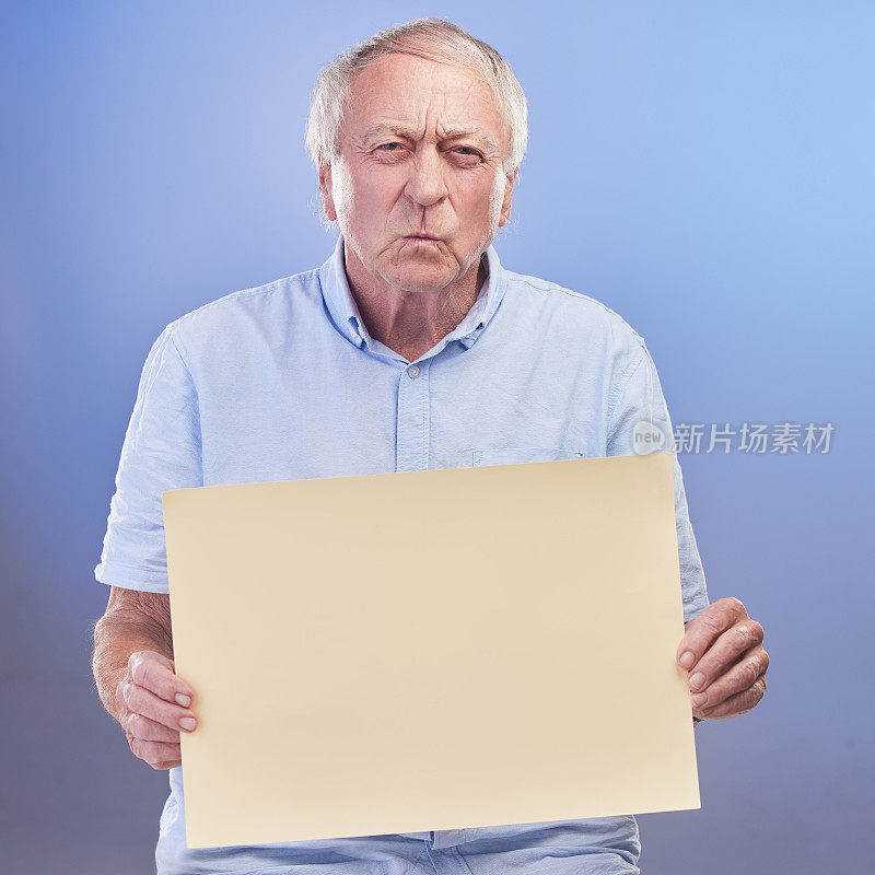 摄影棚拍摄的是一位年长的男子拿着一块空白的牌子，在蓝色的背景下看起来很不开心
