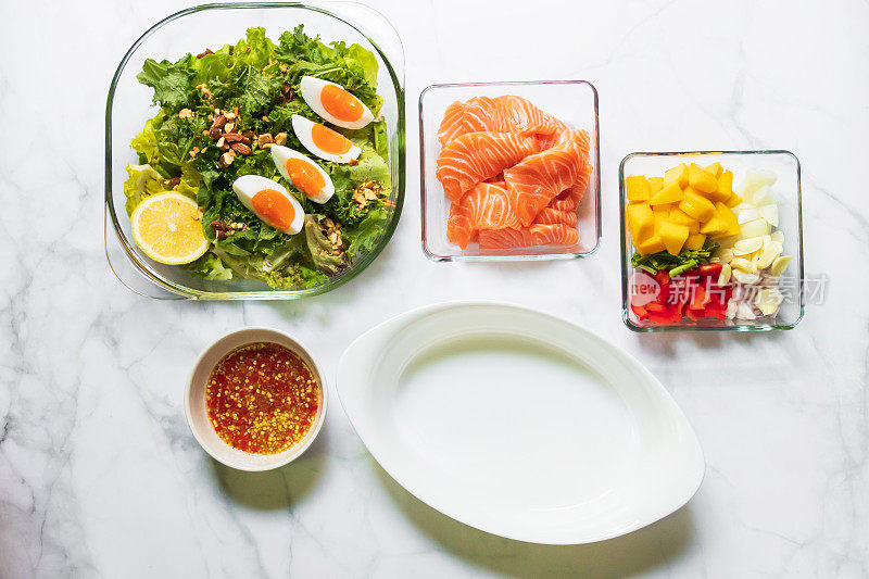 青菜沙拉、羽衣甘蓝、煮鸡蛋、椭圆形白碗，配上美味的辣鲑鱼芒果沙拉配料。白色桌子上的俯视图。