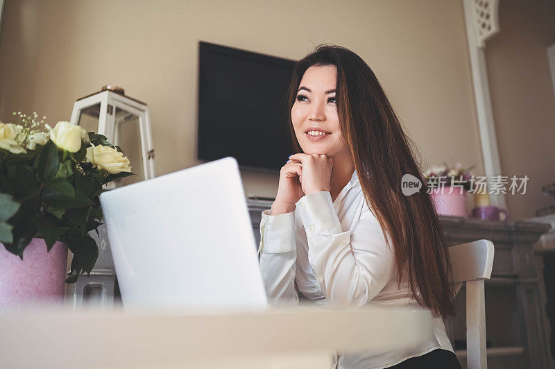 年轻美丽的亚洲商业妇女工作与笔记本电脑放在家里办公室的桌子在线。微笑的自由职业者女士坐在客厅里，拿着白玫瑰花远程工作。