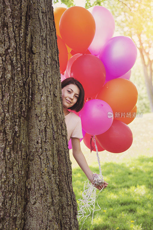 一个女孩拿着一束粉红色的气球躲在树后面。