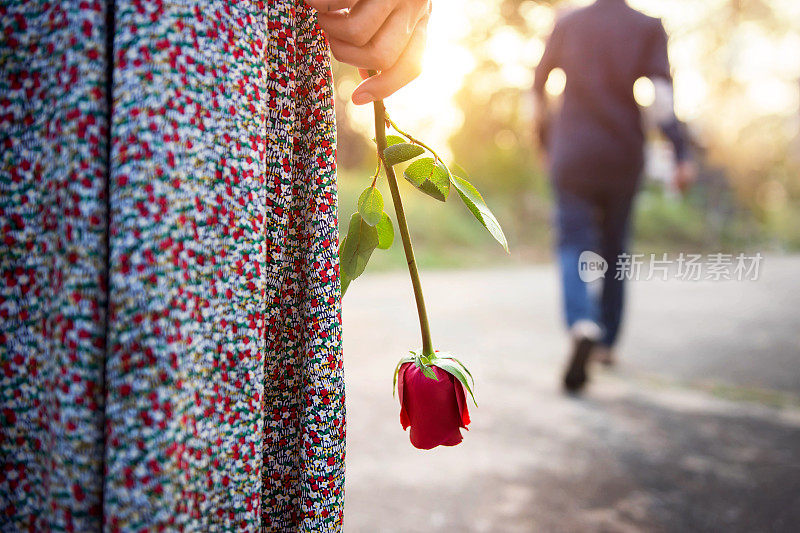 悲伤的爱情在结束的关系概念，心碎的女人站在一只红玫瑰的手，模糊的男人在背后走开作为背景