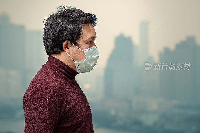 亚洲男子戴着口罩在高公寓的阳台上防止空气污染，可以看到污染和浓雾笼罩曼谷城市景观背景，保健概念