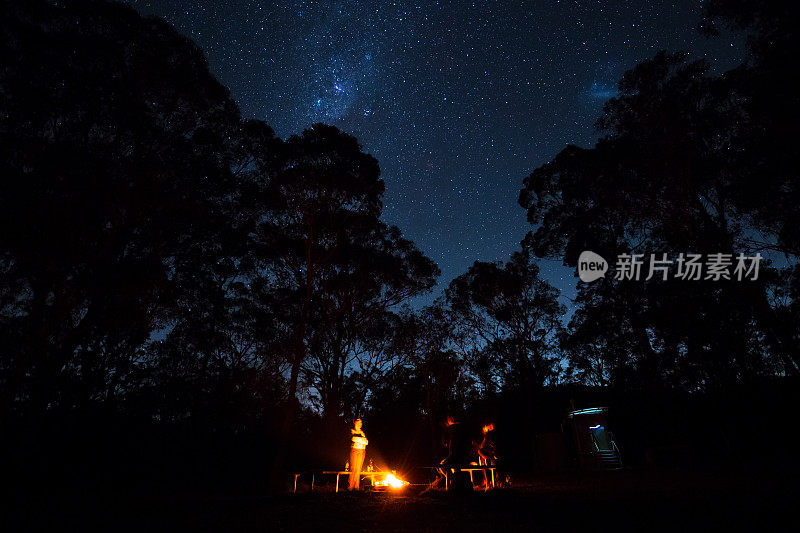 一群露营者围着篝火站着，上面闪烁着明亮的星星