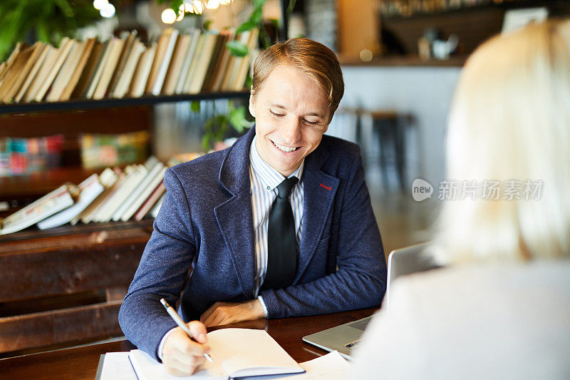 开朗兴奋的年轻商人穿着时髦的休闲夹克，坐在桌子上做笔记，与商业伙伴在图书馆交谈