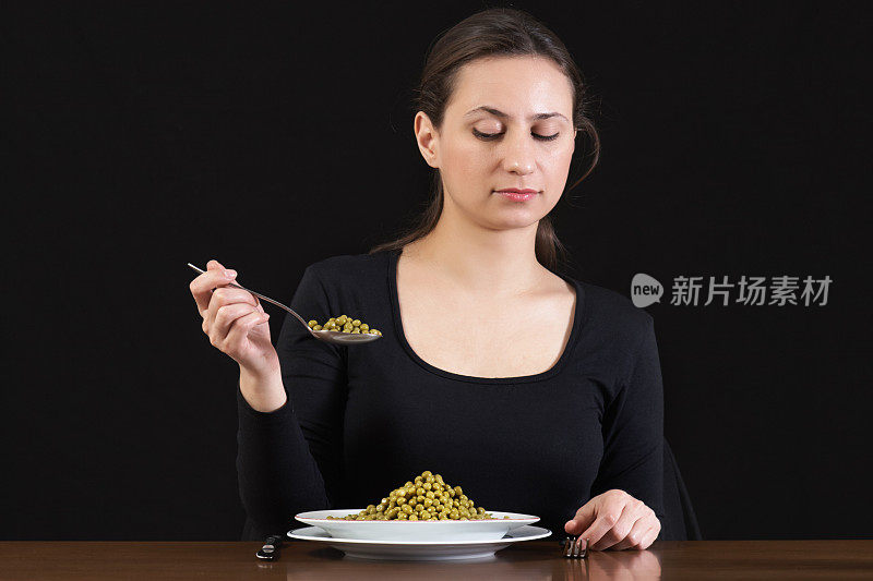 节食。一个年轻的女人拿着满勺绿豌豆坐在桌子上，盘子里有一顿丰盛的减肥餐