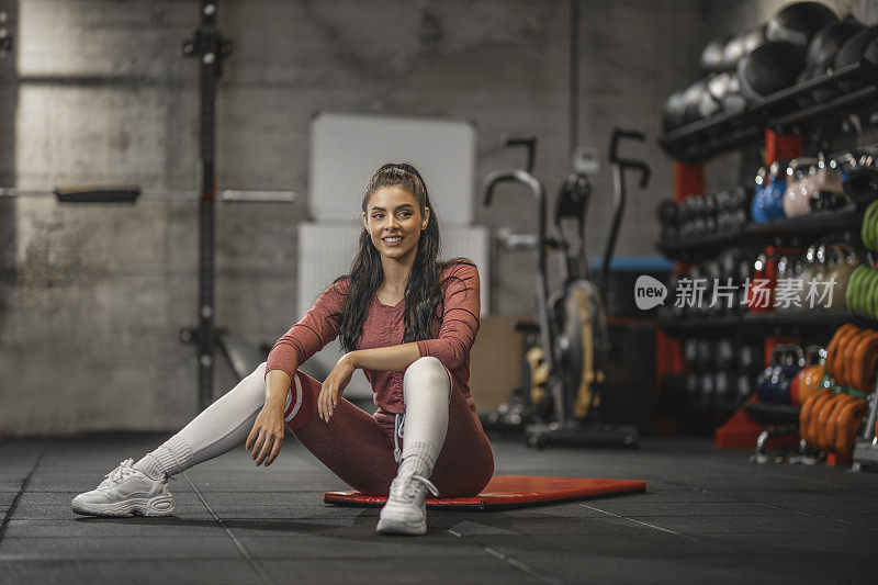 肖像的运动妇女微笑和坐在运动垫在健身房