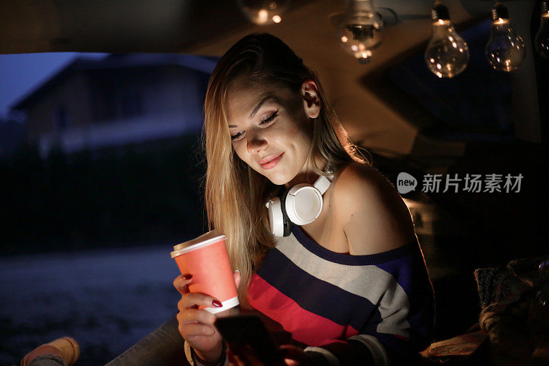 一个年轻漂亮的女人露营在车的后面，放松，同时使用她的手机