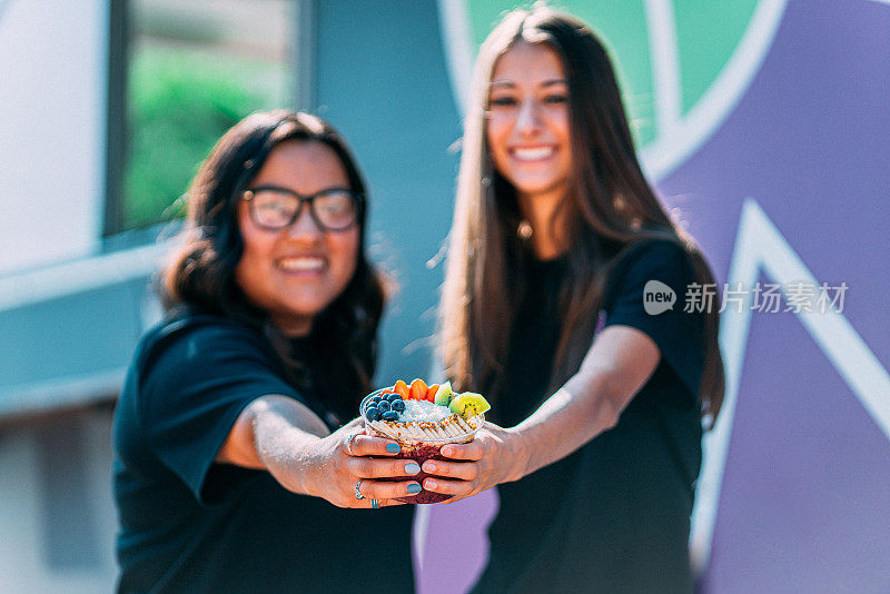 两个年轻的女性朋友站在一起，拿着一个健康的巴西莓食品碗在食品卡车外面