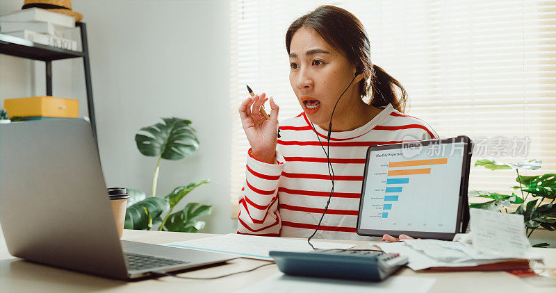 年轻的亚洲女性穿着毛衣，戴着耳机坐在书桌前，用笔记本电脑在线会议，在家里的财务规划师的平板电脑上呈现每月的财务计划。