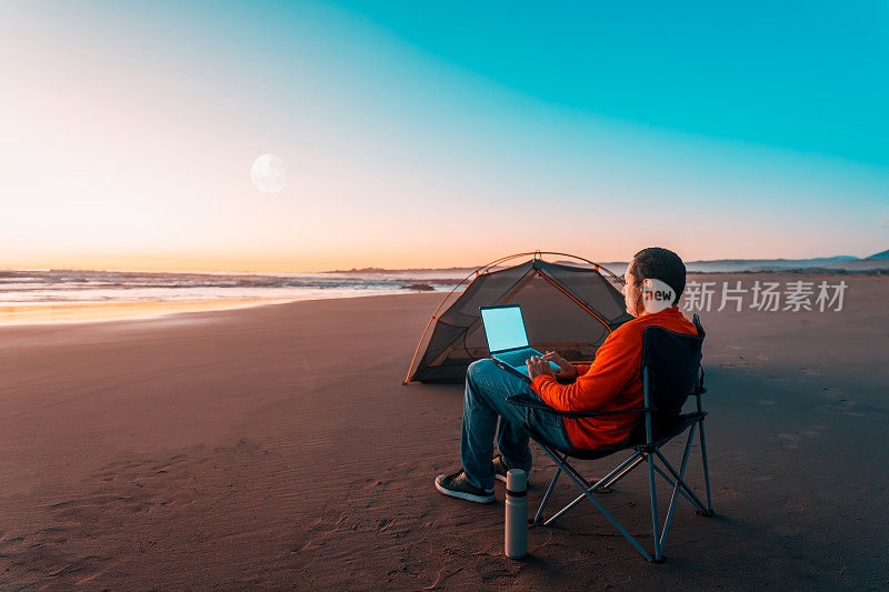 数字游牧民独自坐在海滩岸边，在日落时分用笔记本电脑在帐篷旁工作