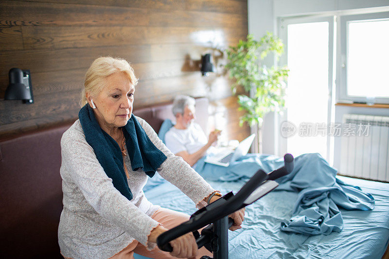 一名老年妇女在室内自行车上锻炼，而她的丈夫在她身后的床上使用笔记本电脑