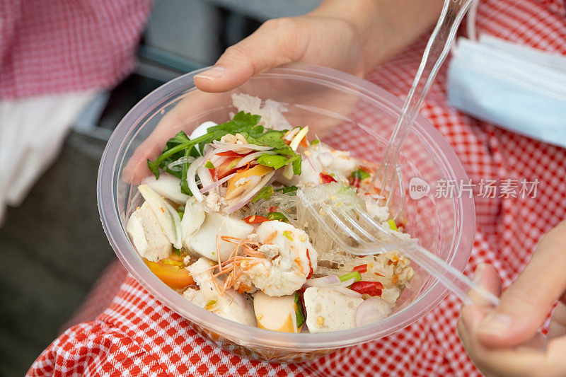 辣粉沙拉配大虾、猪肉香肠、香肠、洋葱和蔬菜，放在女人手里的塑料碗里。