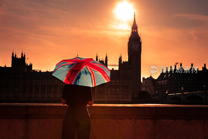夕阳下，一名女子手持英国国旗伞，眺望英国议会大厦和大本钟