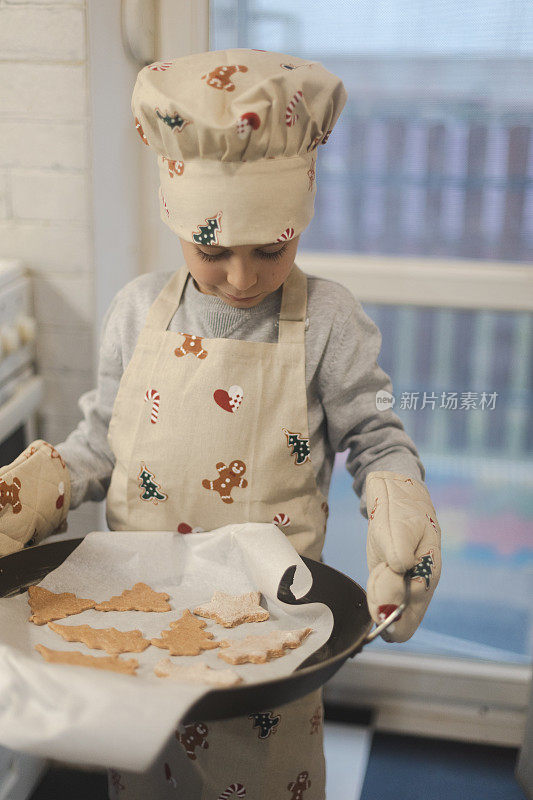 小男孩正在用烤箱手套把装有饼干的托盘从烤箱里拿出来。