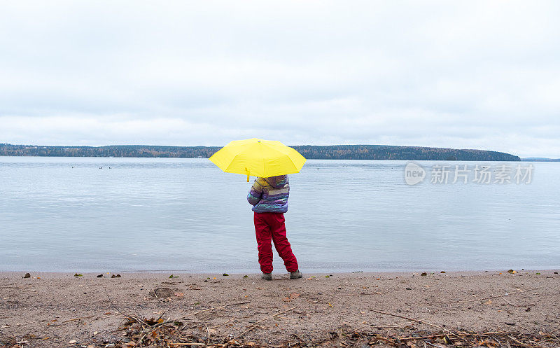 一个8岁的小女孩站在湖边，撑着一把黄色的大伞，欣赏着秋天的风景。后视图。