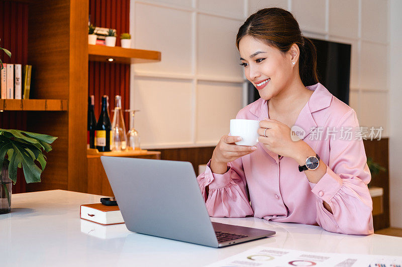 放松的亚洲商务女性自由职业者放松地坐在笔记本电脑前喝咖啡。

在家远程工作或学习，喝杯咖啡，休息一下。