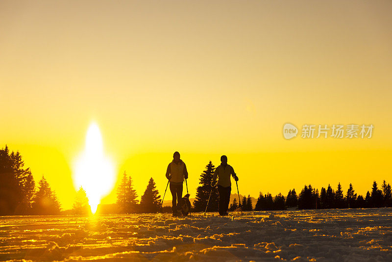 两个穿着保暖衣服的女人拿着滑雪杖，带着伯尔尼山的宠物狗在雪坡上散步