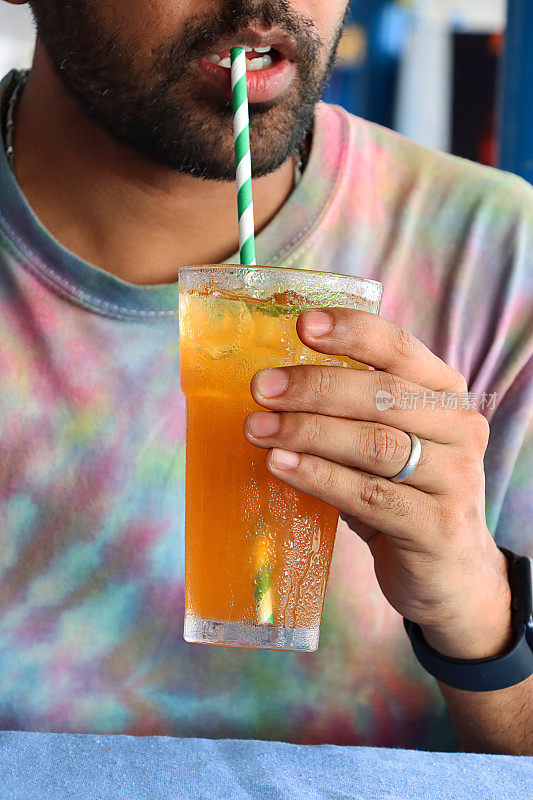 海滩餐厅酒吧场景的特写图片，面面相认的男人用绿色和白色的纸吸管喝着柑橘橙色的无酒精鸡尾酒，冰块和酸橙片，凝结，扎染t恤背景，重点在前景