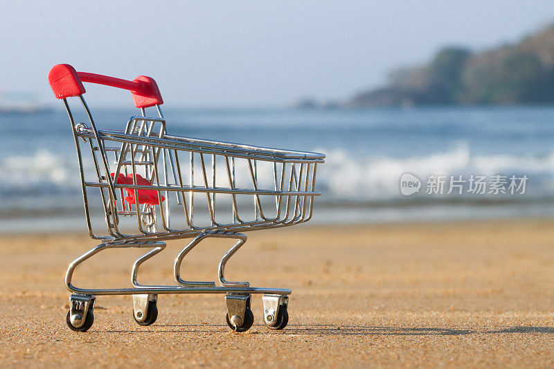 阳光明媚的沙滩上的空的微型购物车特写图像，退潮时的波浪，大海和海岸线，重点放在前景上，暑假购物和旅游的概念，复制空间