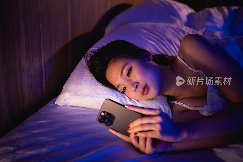 一名亚洲女子躺在卧室的床上玩手机。困倦疲惫，睡不着。失眠，上瘾的概念。女人在黑暗的卧室里用手机刷社交网络。
