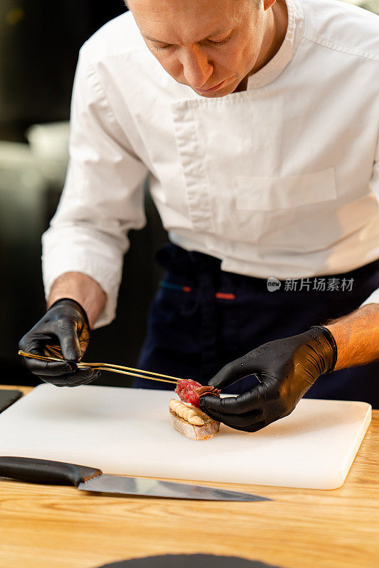 厨师戴着黑色手套的手用镊子夹一块生的腌肉和法棍面包的特写