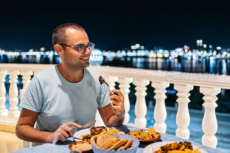 在埃及卢克索，一名年轻男子在餐厅的阳台上享用晚餐，窗外是尼罗河的夜景