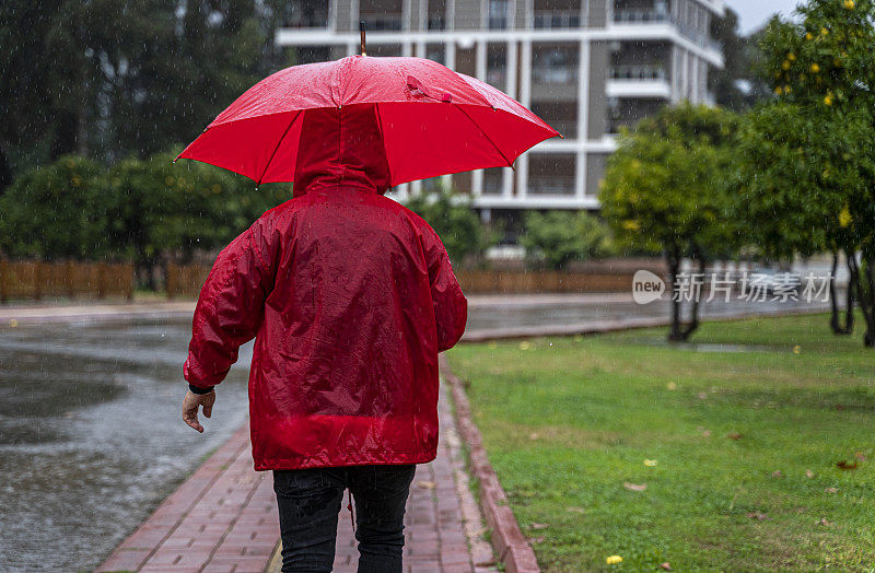 成熟的男人穿着红雨衣，撑着红伞走在雨中的城市街道上