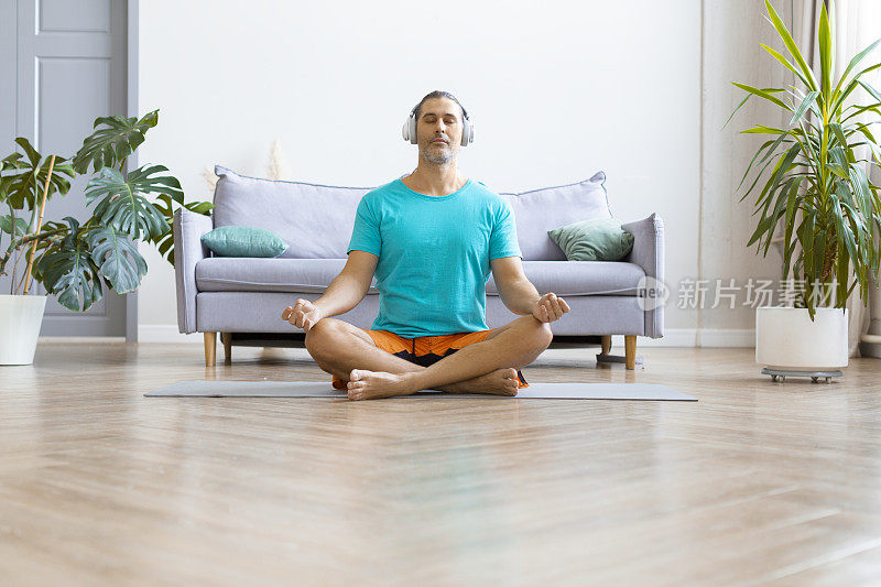 一位中年男子在家练习瑜伽的照片。他以莲花的姿势坐着，戴着耳机听冥想音乐