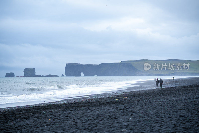 在多云和下雨的天气里，一对夫妇在冰岛南部的黑沙滩上散步