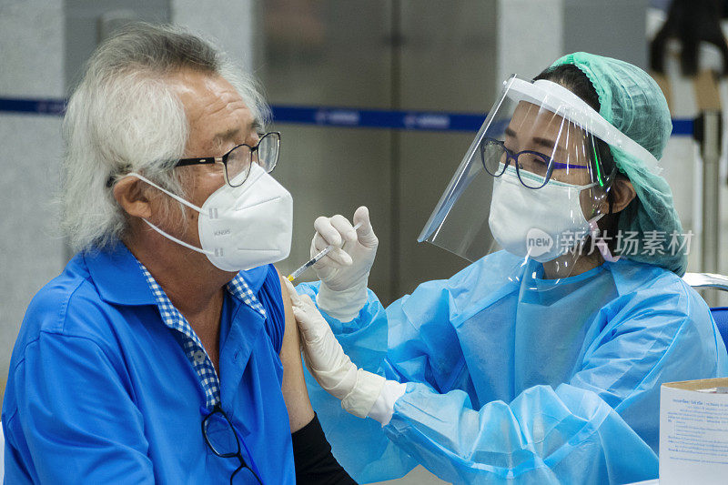 在方苏大车站，一名亚洲医生或护士正在给戴着冠状病毒防护口罩的老年男性患者注射新冠病毒疫苗。