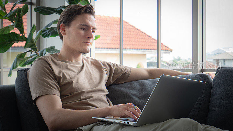 一个英俊的年轻人舒舒服服地坐在长沙发上或笔记本电脑上。一个年轻人坐在工作就像在家里工作或访问社交媒体。