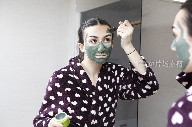 一名年轻女子在家用浴室的镜子前用刷子刷绿色面膜