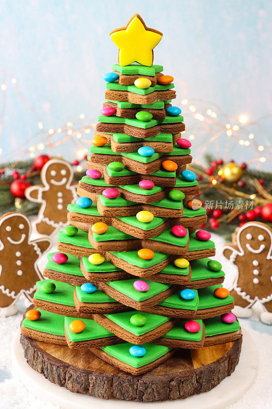近距离拍摄的圣诞树上堆叠着绿色方糖糖霜的姜饼星饼干，装饰着彩色糖果巧克力，上面是黄色的冰星饼干，木制蛋糕架，姜饼人，云杉针，红色浆果