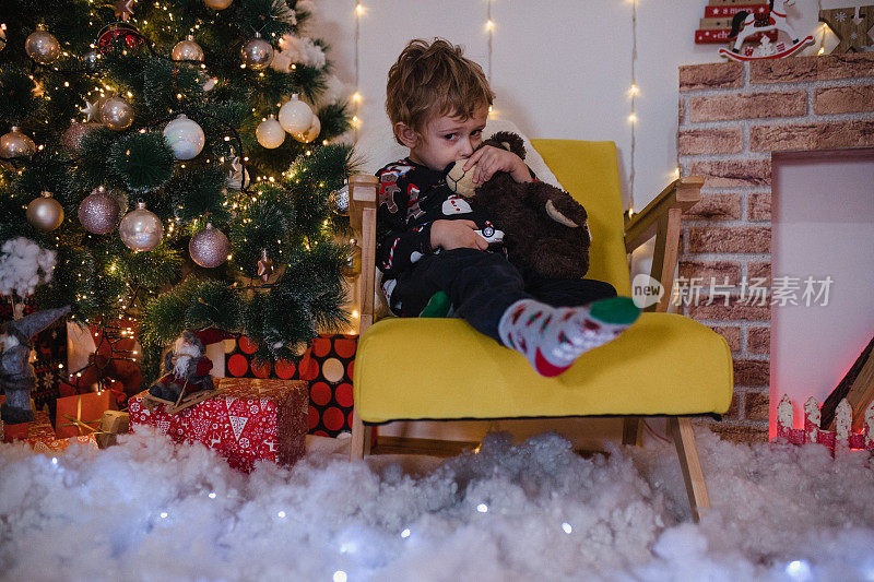 圣诞节那天，快乐的小男孩呆在客厅里。他坐在一棵圣诞树前，抱着她的新玩具熊。